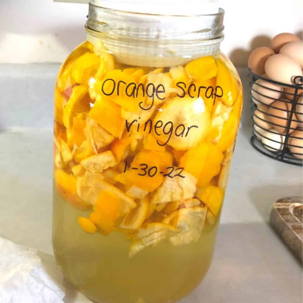 A gallon size glass jar with orange scrap vinegar sitting on the kitchen counter next to a coffee filter and rubber band.