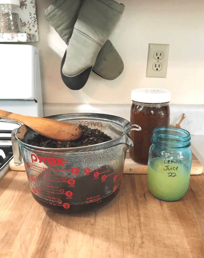A large measuring glass with a metal sieve sitting inside. Herbs are strained through the sieve.  Next to the measuring glass is a jar of honey and a jar of lemon juice.