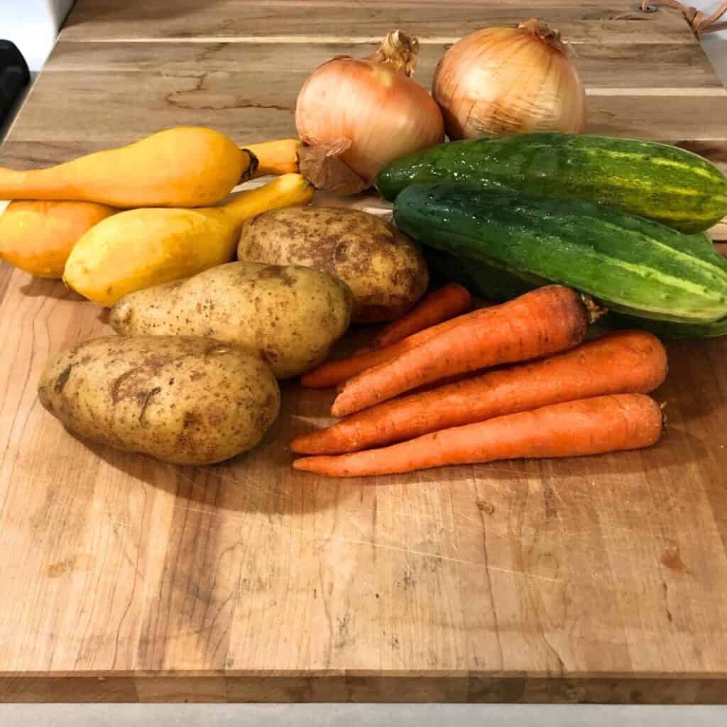 Two onions, two cucumbers, four yellow squash, three potatoes, and six orange carrots sitting on a wooden cutting board.