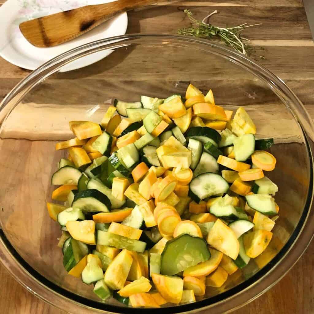 A glass bowl with diced cucumber and yellow squash sitting on a wooden cutting board. A wooden spoon on a white plate and sprigs of thyme tied with kitchen string is next to the bowl.