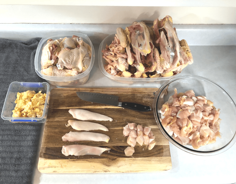 A container of bright orange chicken fat, a container of chicken drumsticks and thighs, a container of chicken carcasses, a glass bowl of one-inch chicken breast chunks, and a wooden cutting board with chicken breasts and a chef's knife.