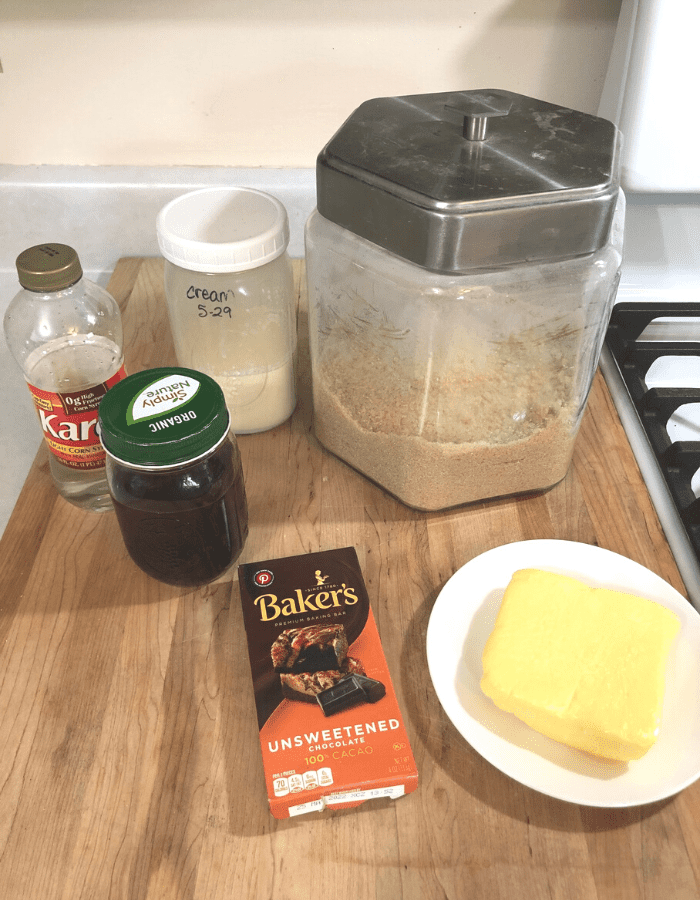 Ingredients for making chocolate fudge including sugar, heavy cream, Karo light corn syrup, vanilla extract, Baker's unsweetened 100% cacao chocolate squares, and butter, sitting on a wooden cutting board.