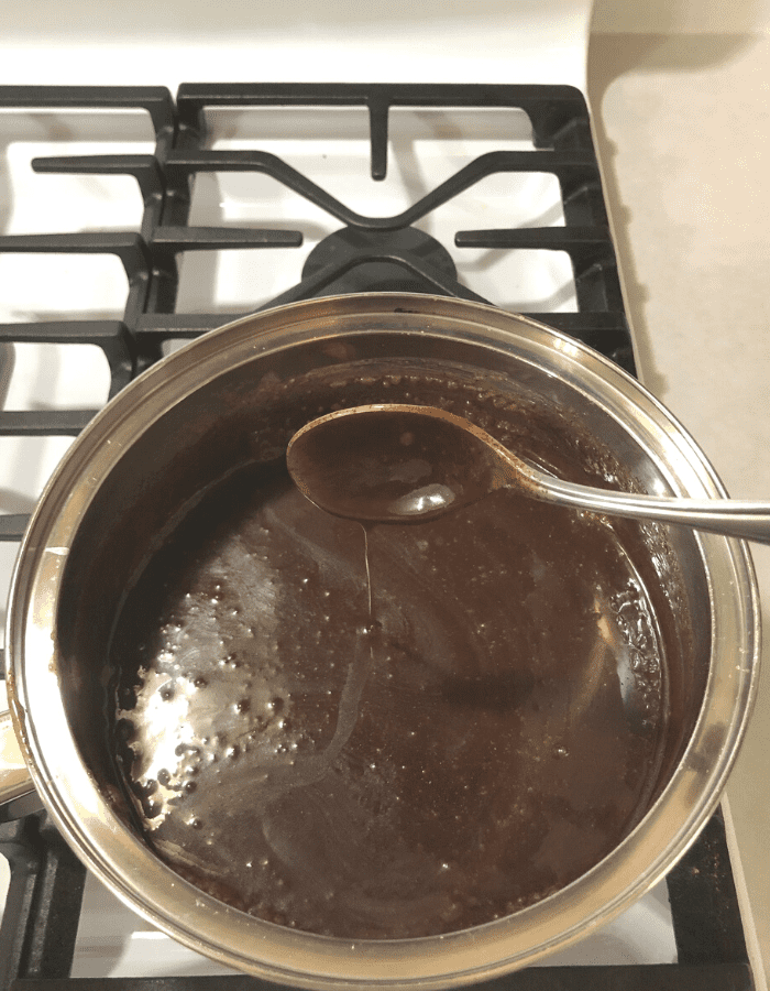 Chocolate fudge in a stainless steel saucepan on the stove while woman stirs with a stainless steel spoon.