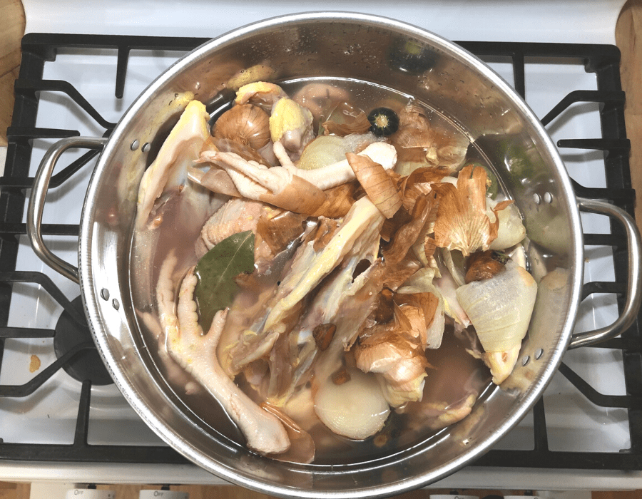 Large stainless steel stockpot on the stove filled with chicken carcasses and scraps, vegetable scraps, and filled with water.