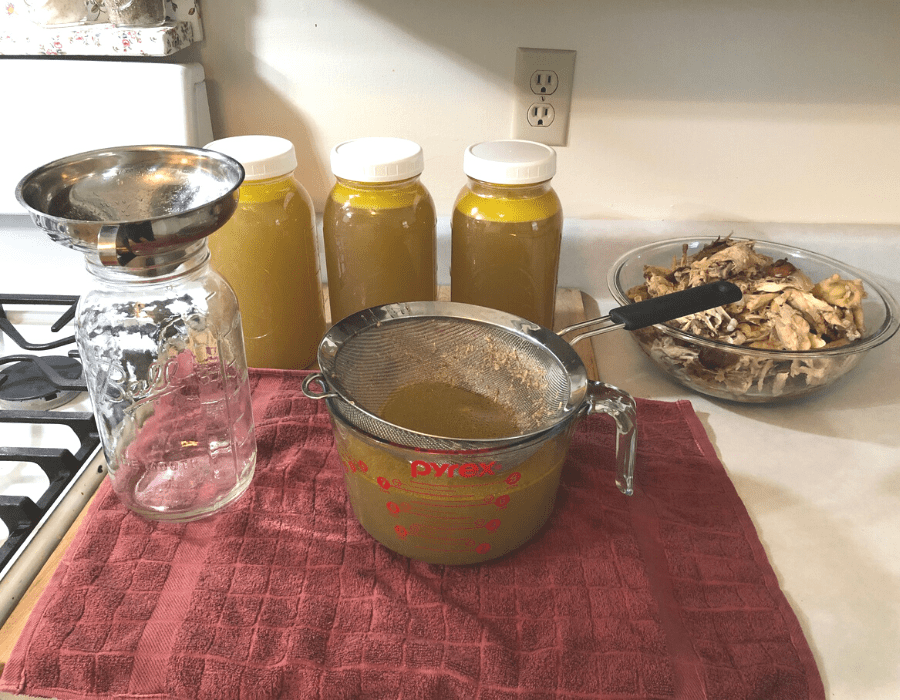 Three half-gallon mason jars and an eight-cup measuring cup filled with stovetop chicken bone broth sitting on a red towel on the kitchen counter. A glass bowl to the side has chicken bone broth scraps. An empty half-gallon mason jar with a stainless steel funnel on top is next to the eight-cup measuring cup with a fine mesh sieve resting on top.