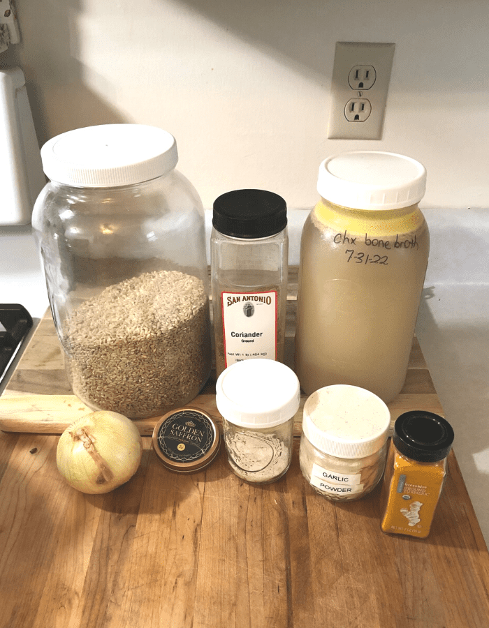 A glass jar of brown rice, a half-gallon mason jar of chicken bone broth, an onion, ground coriander, golden saffron, salt, garlic powder, and ground turmeric on a wooden cutting board on the counter.