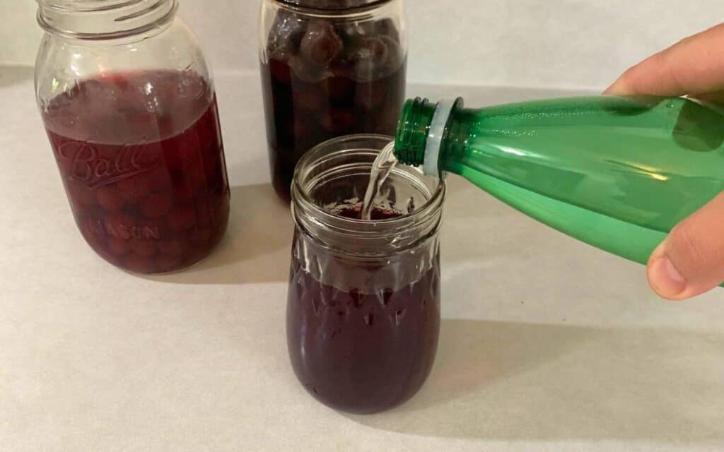 Woman pouring mineral water into a glass jar filled halfway with cranberry juice and cherry juice.