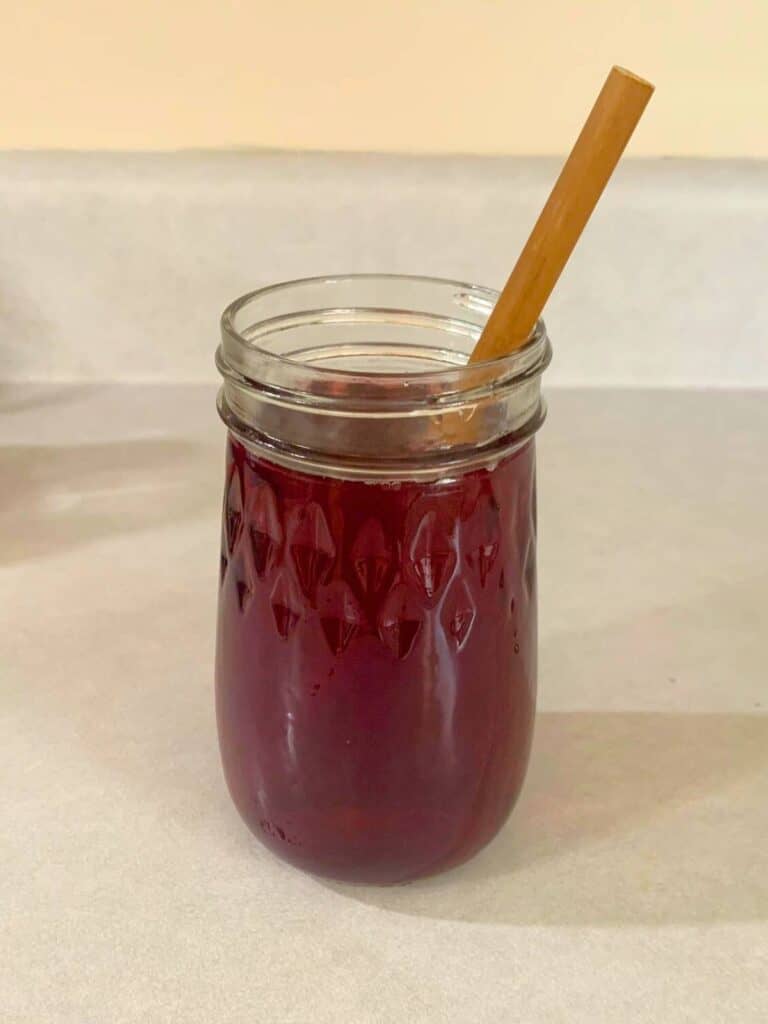 A clear, fluted glass filled with cranberry juice is sitting on a kitchen counter. A wood bamboo straw is set inside.