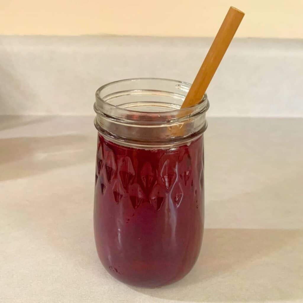 A fluted clear glass filled with cranberry juice is sitting on a kitchen counter. A wooden bamboo straw is set inside the glass.