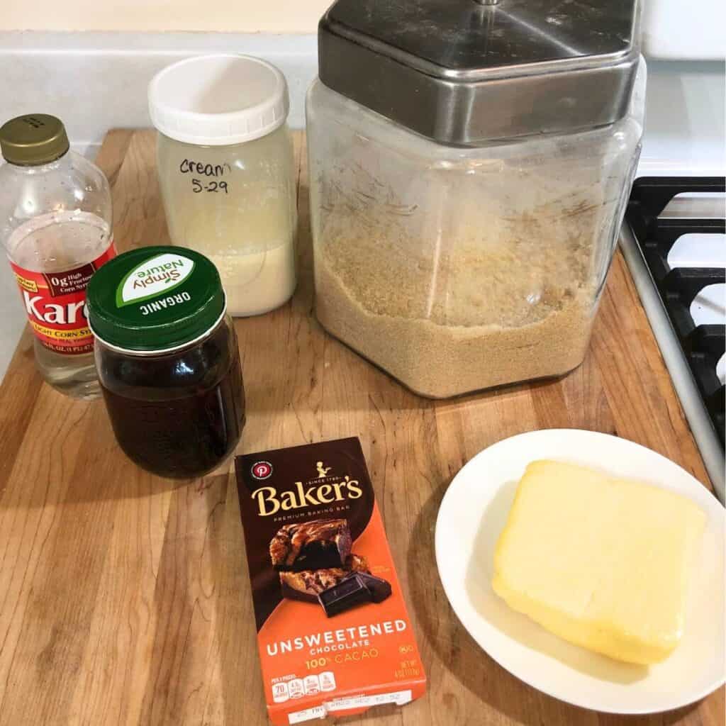 Ingredients for making chocolate fudge sitting on a wood cutting board. Ingredients shown are sugar, unsweetened chocolate, cream, butter, vanilla extract, and corn syrup.