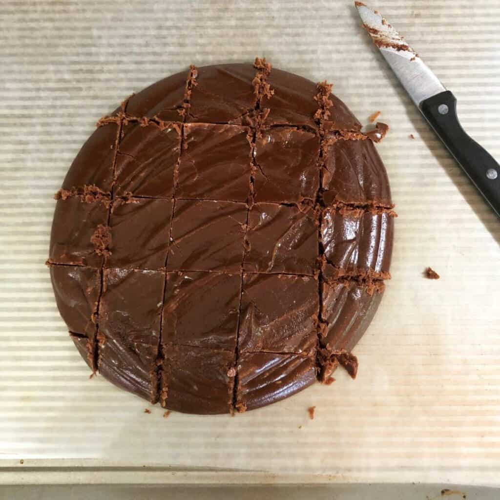 Chocolate fudge poured out into a rough circle shape on wax paper. It has been cut into squares and a sharp knife is laying next to it.