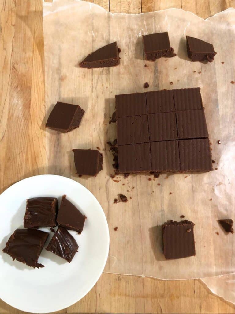 Chocolate fudge pieces on wax paper on a wood countertop sitting next to a small white plate with four pieces of fudge.