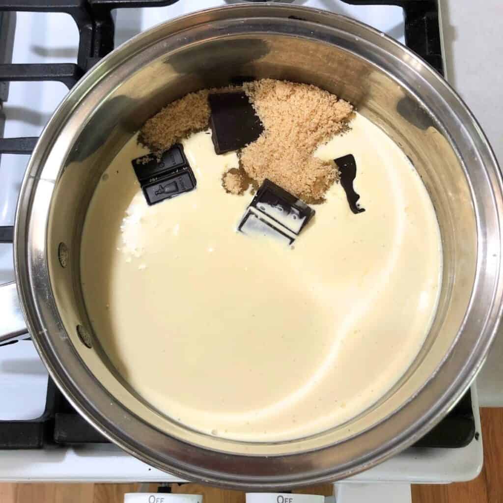 Stainless steel saucepan on a stove with sugar, cream, chocolate, and corn syrup inside.