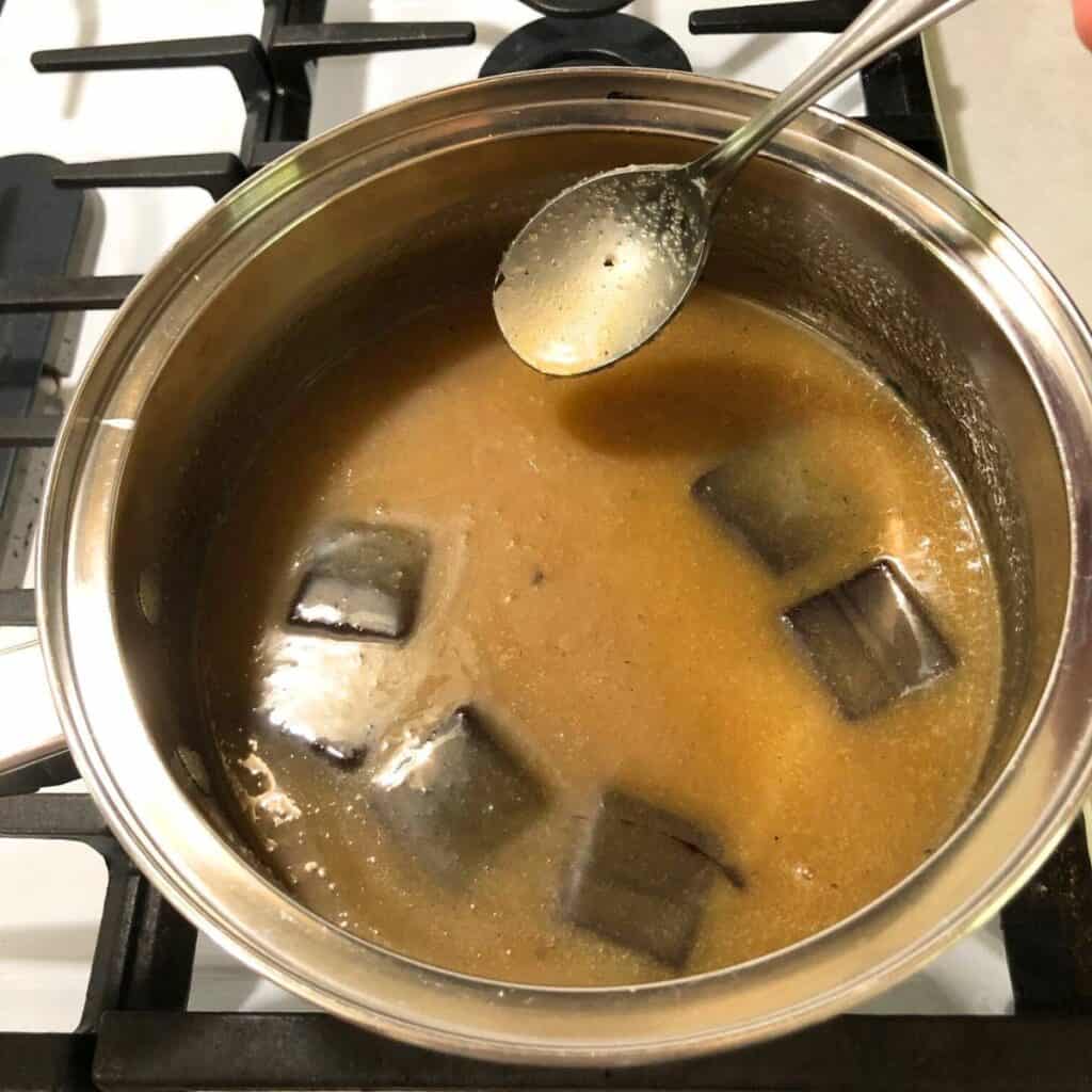 Woman stirring chocolate fudge ingredients in a stainless steel saucepan on a white gas stove.