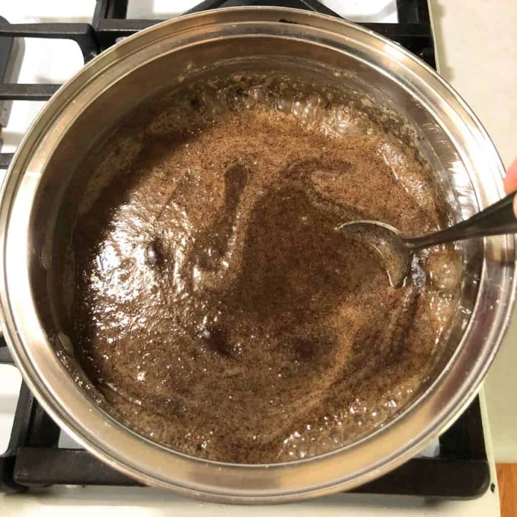 Woman stirring chocolate fudge ingredients in a stainless steel saucepan on a white gas stove. It is bubbling in the pot.