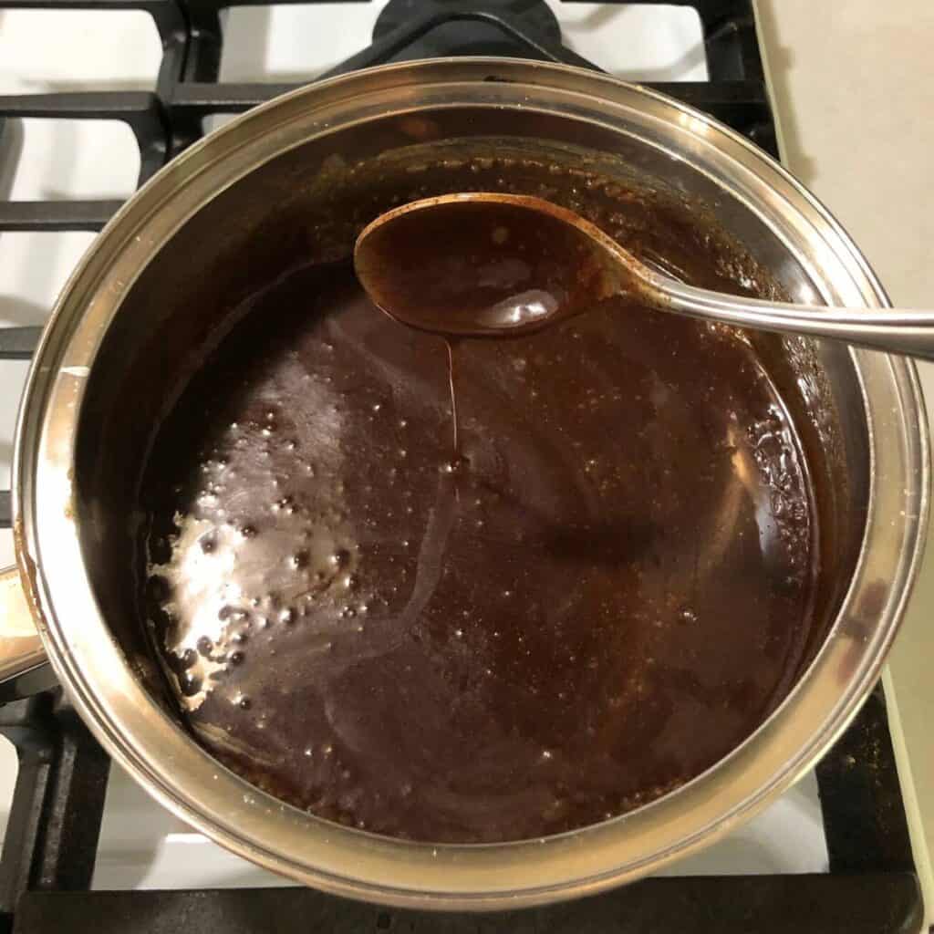 Woman holding a stainless steel spoon that has been dipped into hot chocolate fudge. She is holding the spoon up to show the fudge coating the spoon and drizzling back down into the pot.