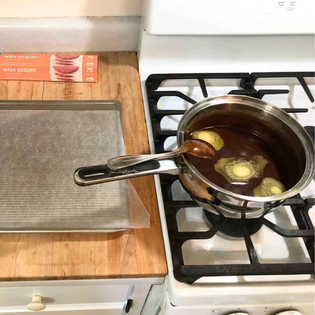 Stainless steel saucepan of chocolate fudge with three tablespoons of butter added to it, sitting next to a cookie sheet with wax paper on top of a wooden countertop.