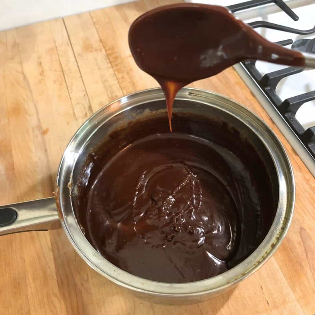 Woman stirring vanilla extract into chocolate fudge in a stainless steel saucepan. She's showing how the fudge is sticking to the spoon and dripping in ribbons back into the pot.