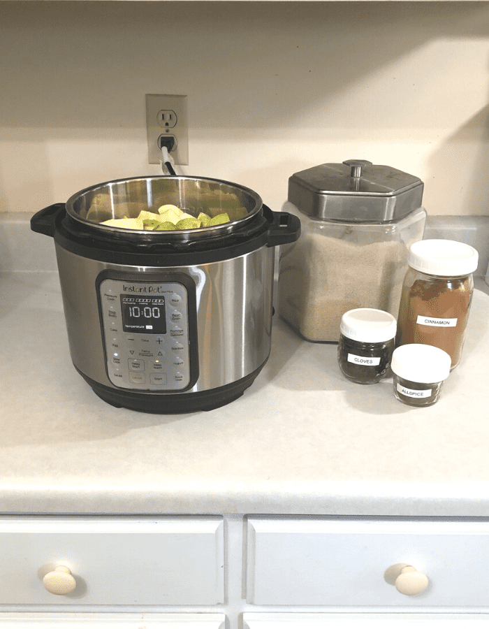 An instant pot with cut apples filling it, a glass canister of organic sugar, and glass mason jars of ceylon cinnamon, ground cloves, and ground allspice sitting on the kitchen counter.
