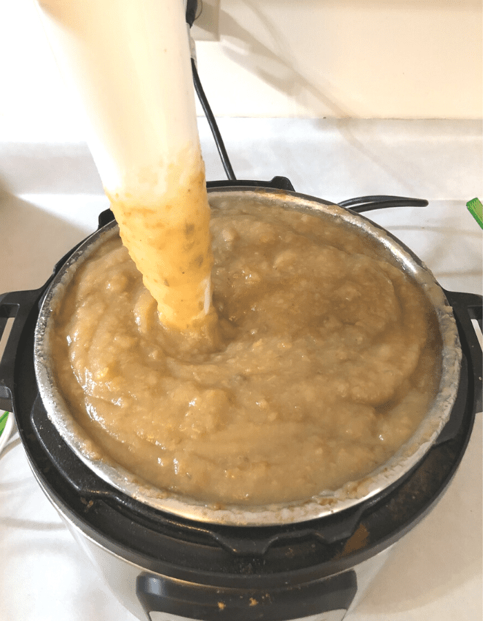 An instant pot filled with apple butter has a white an immersion blender inside blending it up.
