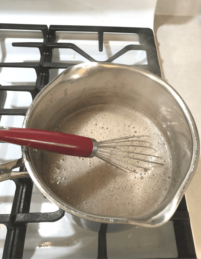 A stainless steel saucepan with four cups of apple juice, one-fourth cup of lemon juice, four teaspoons of calcium water, and sugar and pectin mixed in with a red whisk.