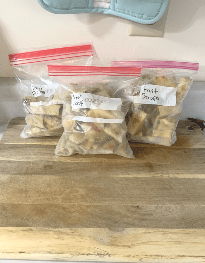 Three quart-size freezer bags of frozen apple scraps such as apple cores, sitting on a wooden cutting board on the counter.