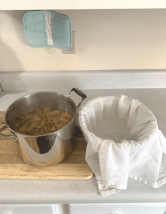 A silver pot of hot apple scraps and apple juice sitting on a wooden cutting board next to a bowl lined with a white flour sack towel.