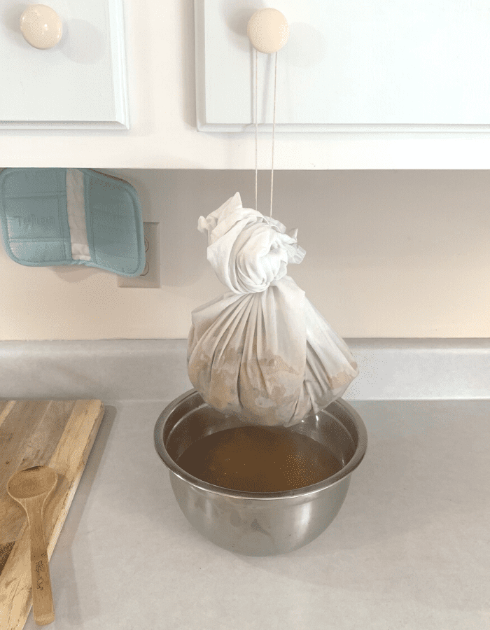 A white flour sack towel filled with hot apple scraps and apple juice hanging from an upper cabinet knob over a silver bowl. Apple juice is dripping into the bowl underneath the bag of apples.