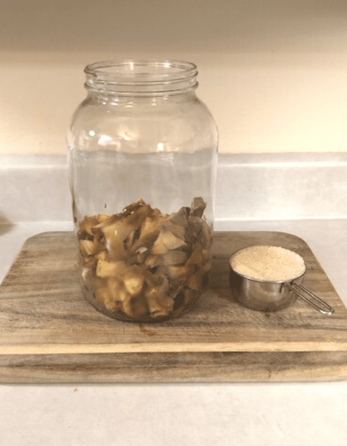 A glass jar filled half-way with apple scraps, sitting on a wooden cutting board on the counter next to a measuring cup of sugar.