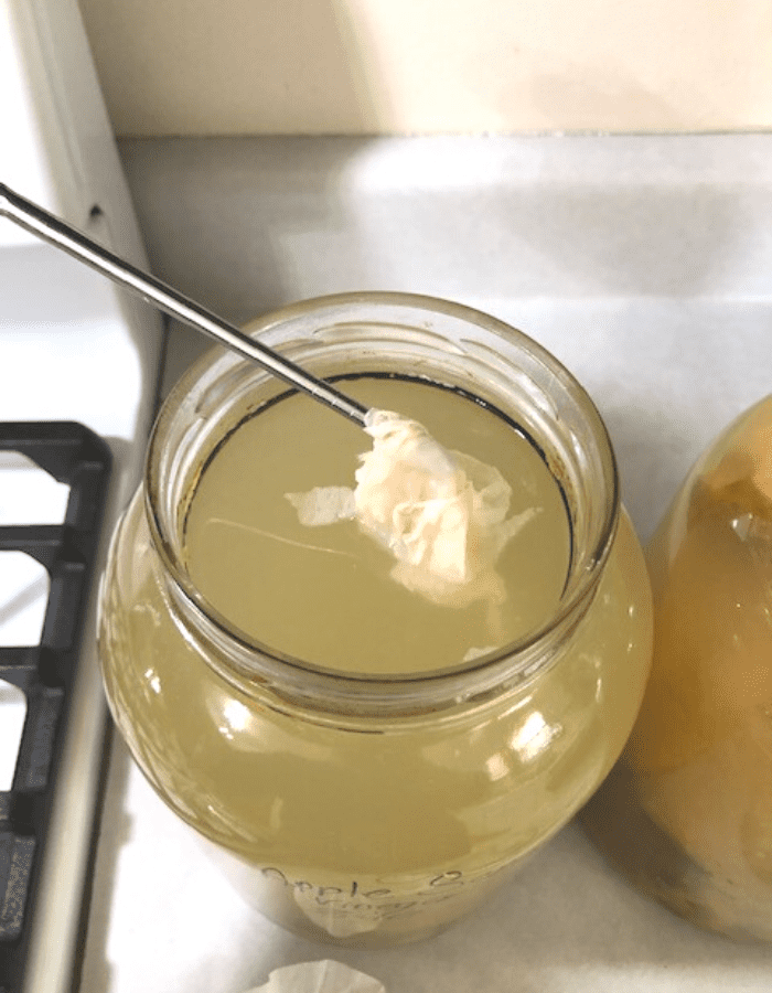 A glass jar of apple scrap vinegar sitting on the counter with a stainless steel chopstick stirring it.