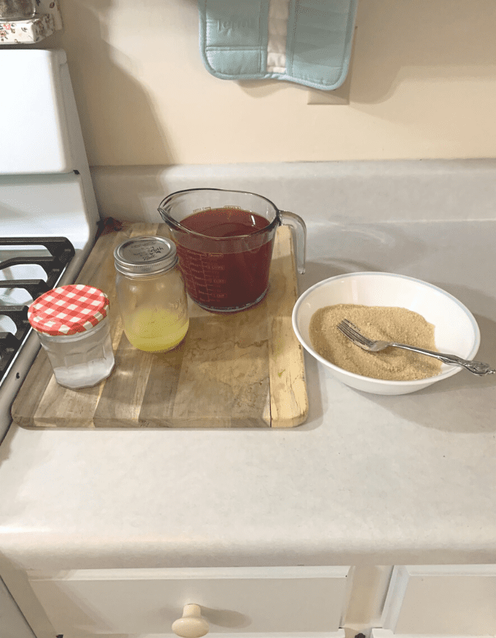 A measuring cup with cranapple juice, a mason jar of lemon juice, another mason jar of Pomona's Pectin calcium powder in water all sitting on a wooden cutting board on a counter.  Next to it is a white bowl of sugar mixed with Pomona's pectin powder.  A fork is sitting in the bowl.