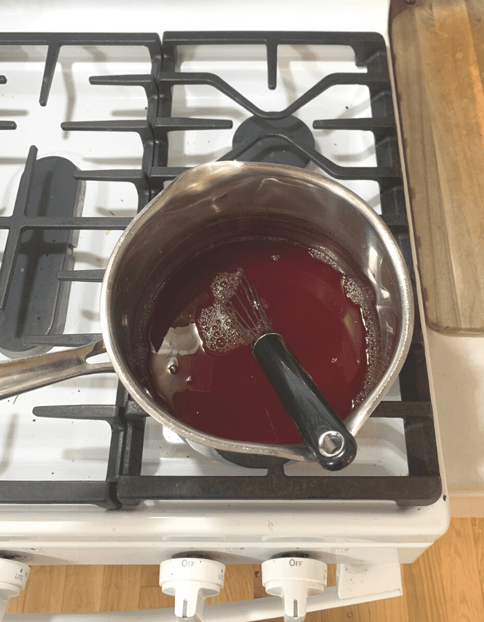 A medium saucepan on the stove with liquid cranapple jelly inside. A wire whisk is sitting in the pot.