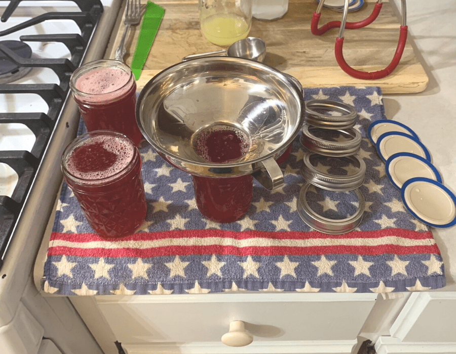 Half-pint jelly jars with cranapple jelly inside and a stainless steel funnel in one. Canning rings and canning lids are sitting next to the jars on a towel.