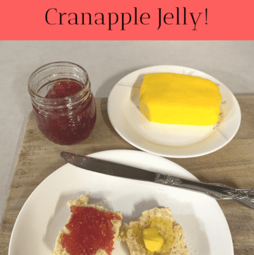 A mason jar of cranapple jelly sitting next to homemade butter on a white plate and another white plate with a biscuit cut in half. Butter is melting on one half and cranapple butter is spread on the other half. The plates and mason jar are sitting on a wooden cutting board on the counter. A knife is resting on the biscuit plate.