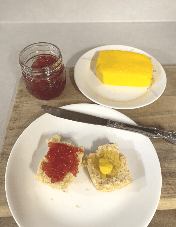 A mason jar of cranapple jelly sitting next to homemade butter on a white plate and another white plate with a biscuit cut in half. Butter is melting on one half and cranapple butter is spread on the other half. The plates and mason jar are sitting on a wooden cutting board on the counter. A knife is resting on the biscuit plate.