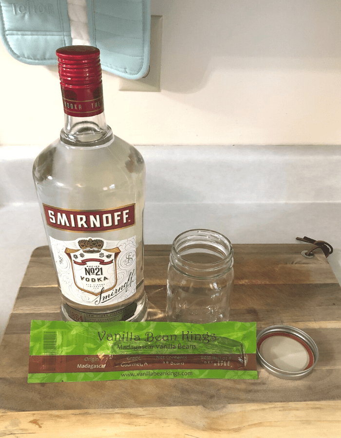 A bottle of vodka, a package of Madagascar vanilla beans, and a glass pint jar with lid sitting on a wooden cutting board on the counter.