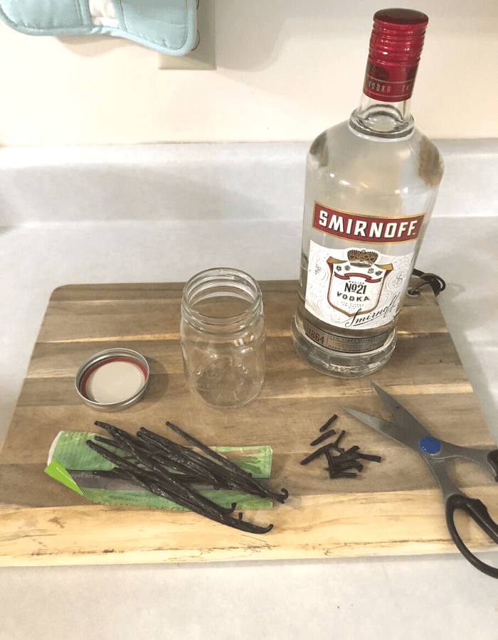 A glass bottle of vodka, ten Madagascar vanilla beans, some cut in half and then in segments of one-inch, an empty glass pint jar with lid, and scissors sitting on a wooden cutting board on the counter.