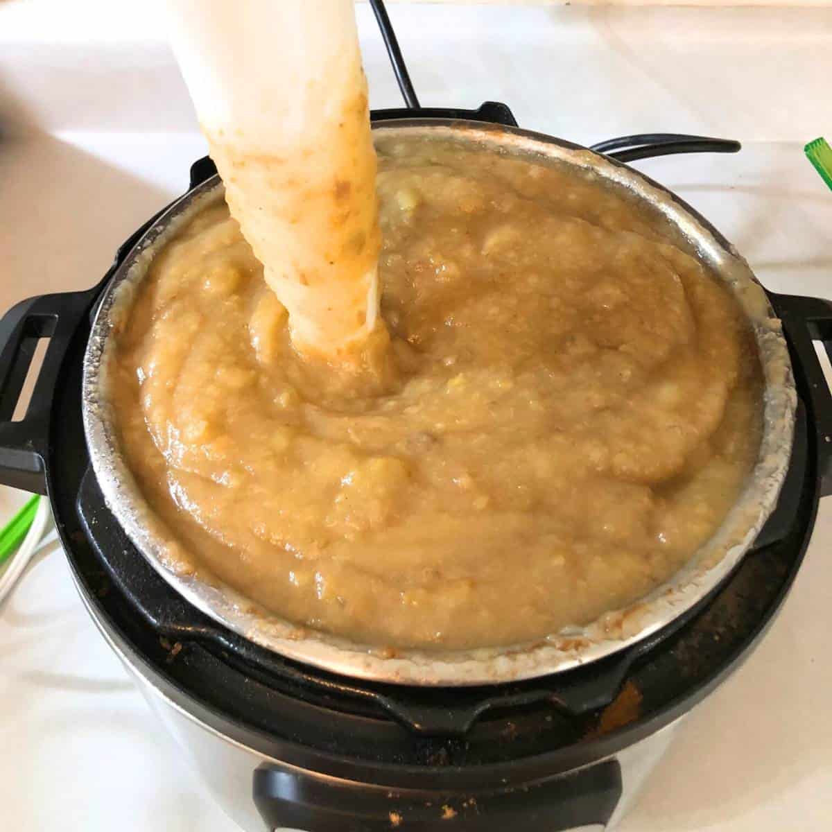 An instant pot filled with apple butter has a white an immersion blender inside blending it up.