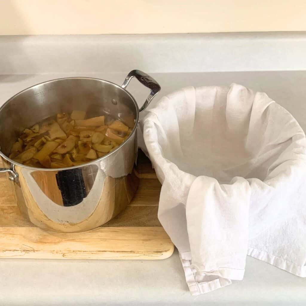 A large pot of steaming apple cores and water on a wooden cutting board next to a bowl with a white flour sack towel draped inside.