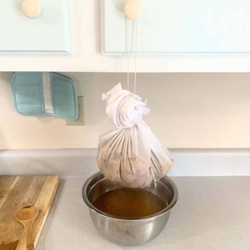 A white flour sack towel made into a bag with a kitchen string slip knot and steamed apple cores are inside the bag and dripping apple juice into a bowl underneath.