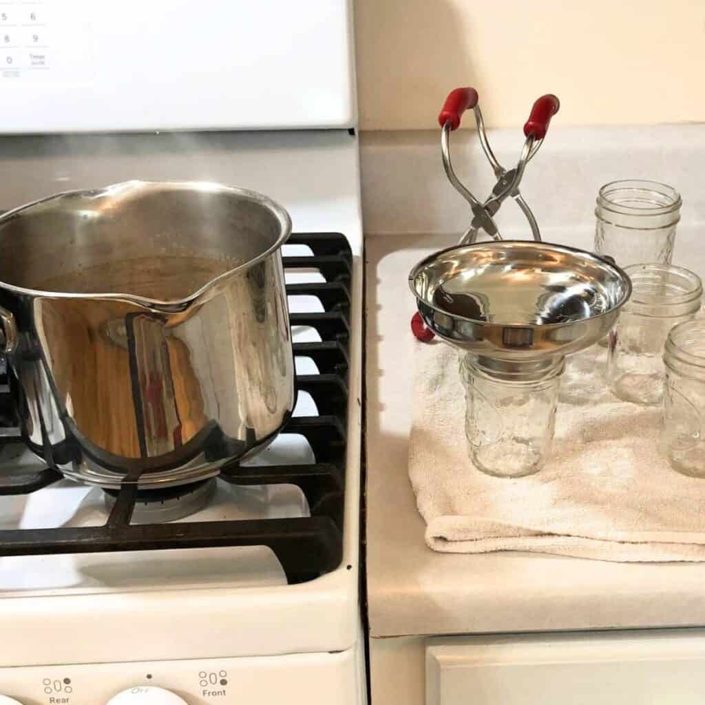 Large stainless steel pot on a stove with several empty mason jars on the counter next to it along with a canning funnel and jar lifter.