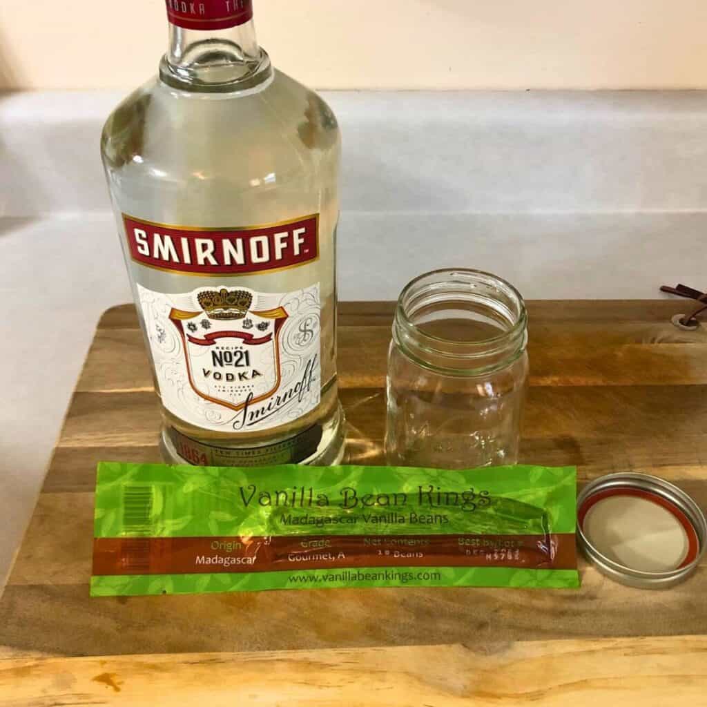 A glass bottle of vodka sitting on a wood cutting board next to an empty mason jar and a package of vanilla beans.