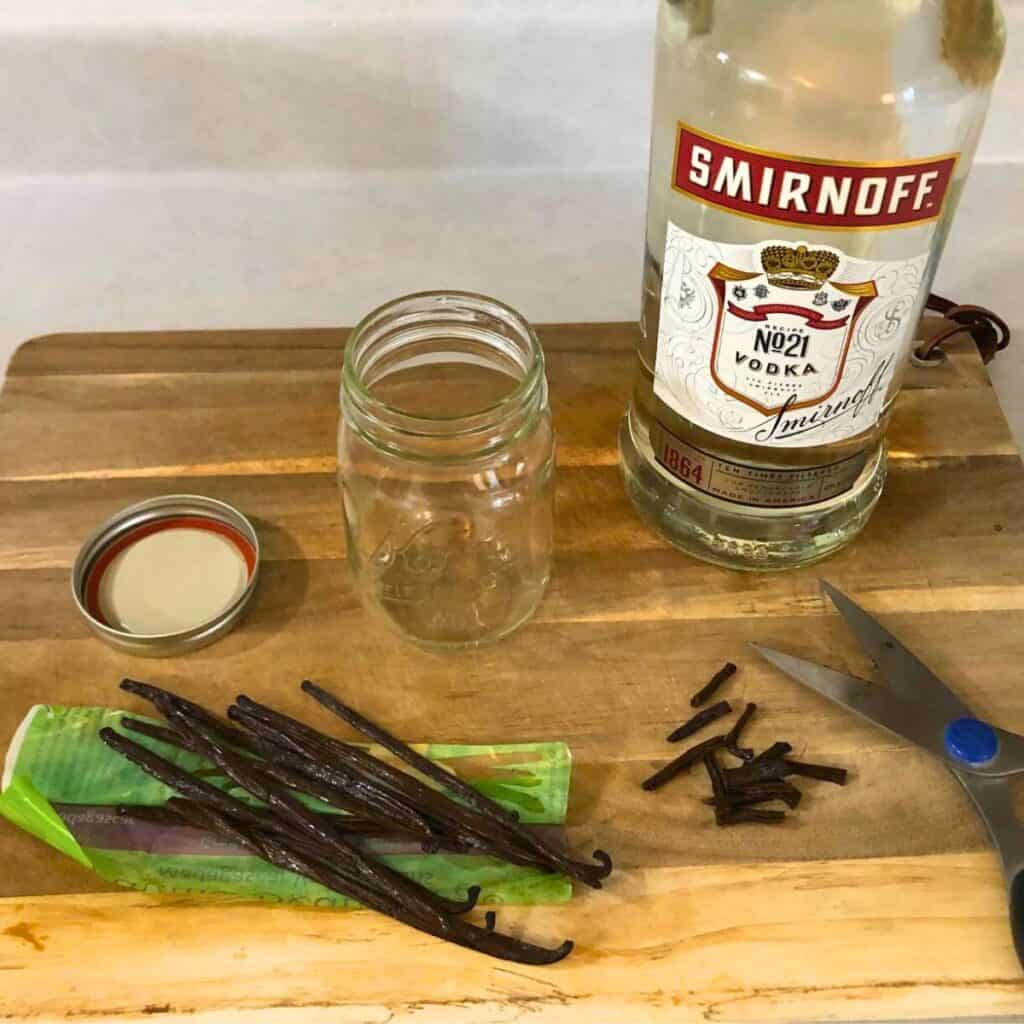 A bottle of Smirnoff vodka sitting on a wood cutting board next to vanilla beans that have been cut and an empty mason jar.