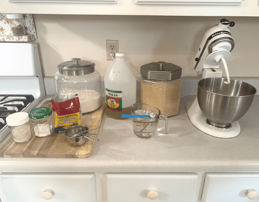 A kitchen counter with ingredients and equipment for making easy sandwich bread. Included are a stand mixer with dough hook attachment, measuring cups, measuring spoons, a wooden cutting board, and a hot liquid thermometer. Ingredients out include a glass canister of flour, a glass canister of organic sugar, a mason jar of salt, instand dry year, olive oil, and two cups of water in a glass measuring cup.