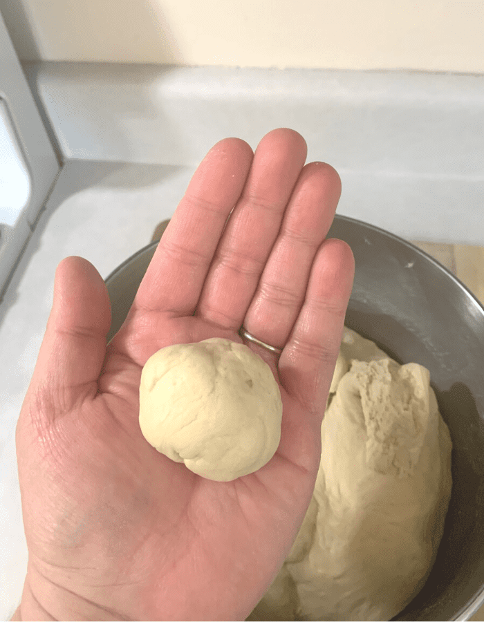 A woman's hand holding a golf ball size piece of bread dough, rolled into a ball. 