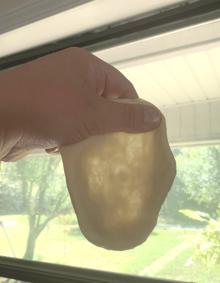 A woman holding up a flattened piece of dough in front of a window and light is showing through the center of the dough.