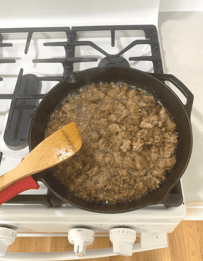 Two pounds of sausage frying in a cast iron skillet on a stove with a wooden spoon resting  on the handle.