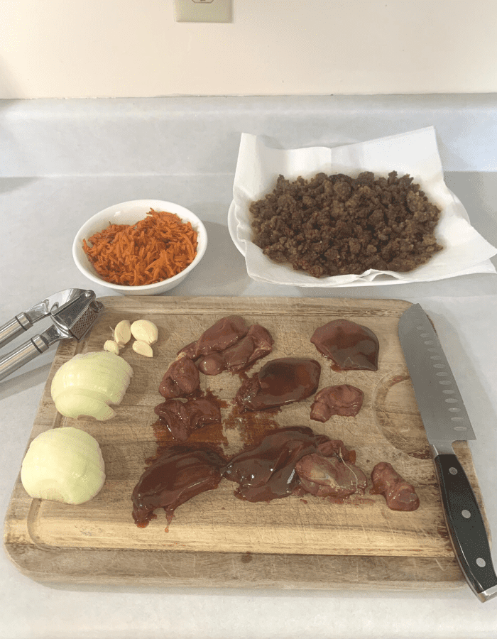 A wooden cutting board with sliced onion, four cloves of garlic next to a garlic press, and liver next to a chef's knife. The wooden cutting board is next to a white bowl filled with grated carrot and a platter of sausage draining on paper towels.