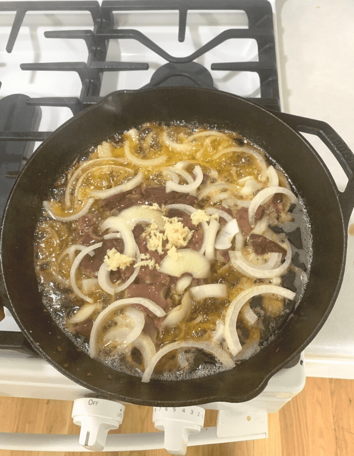 Chopped liver, sliced onion, and minced garlic in a cast iron skillet with sausage fat frying on the stove.