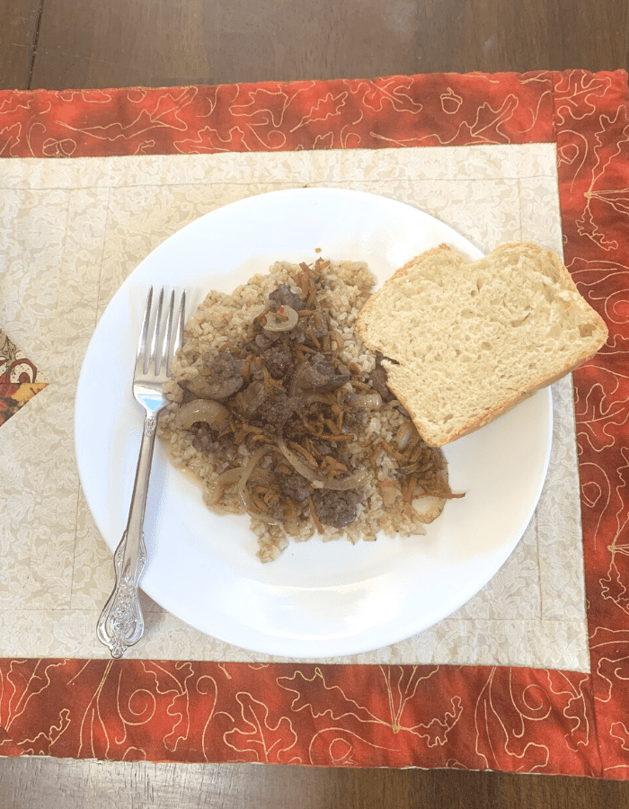 Sausage and liver recipe over a bed of brown rice with a slice of bread and a silver fork on a white plate. The plate is on a fall themed quilted place mat.